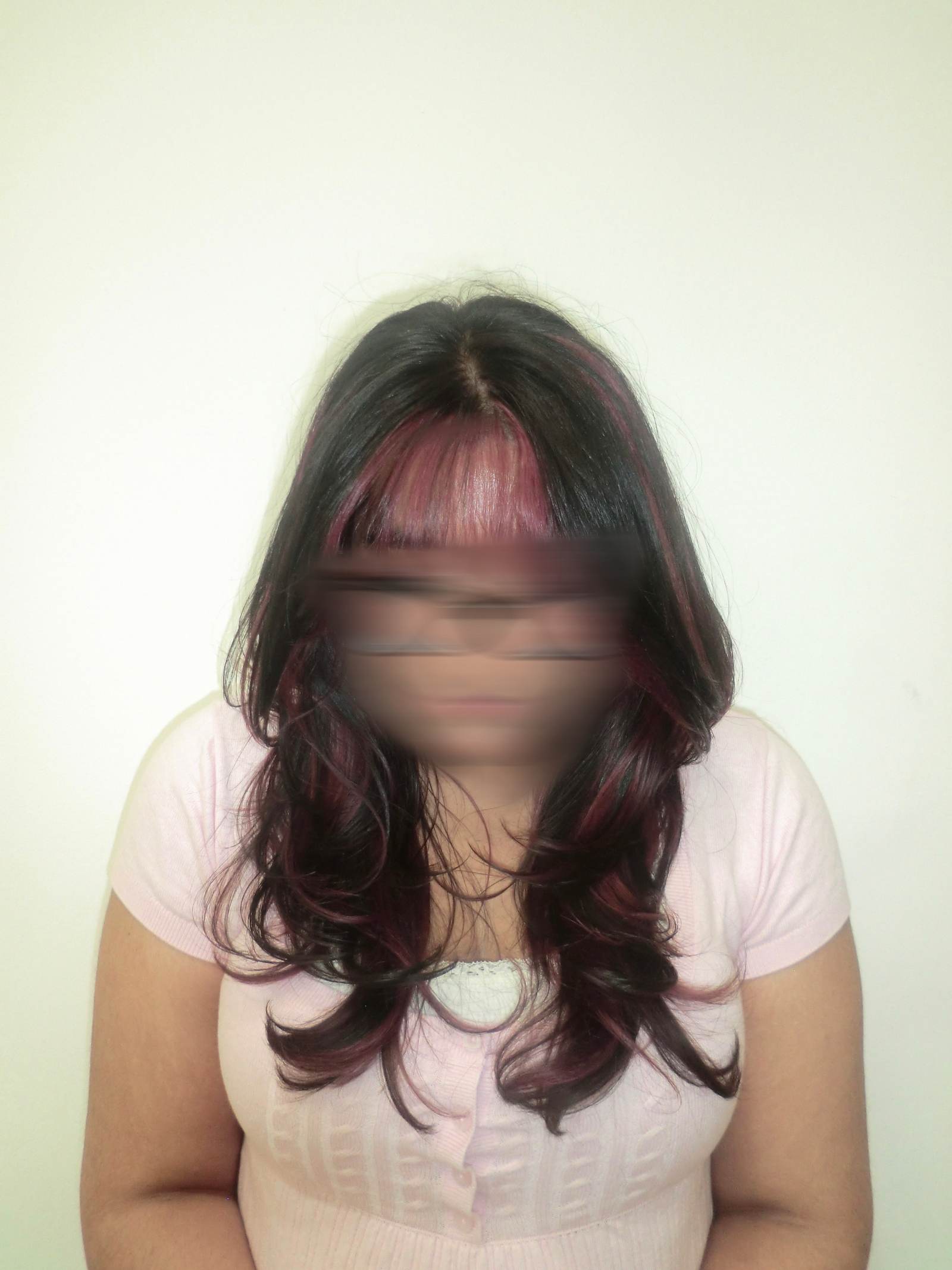A front profile of a person with long, wavy black hair featuring a high-contrast pink transformation. The full, straight-across bangs are dyed a vibrant magenta pink, which continues into "peekaboo" sections underneath the black top layers. The pink strands frame the face and peek through the dark curls, creating a bold and edgy two-tone look.