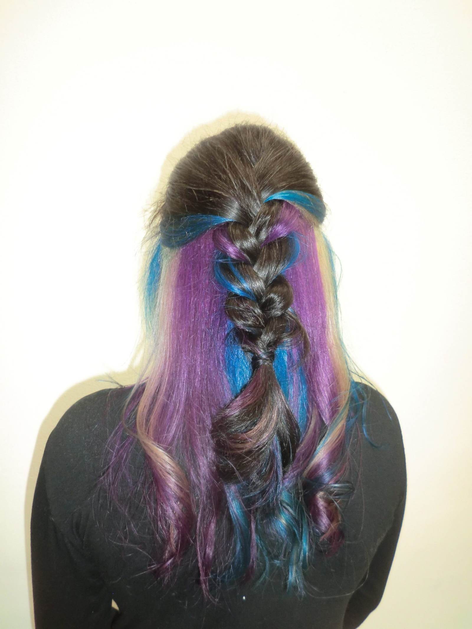 A close-up, top-down view of a thick French braid that reveals vibrant layers of electric blue and bright purple hair underneath a dark brunette surface. The colorful strands are woven into the braid, creating a striking contrast.