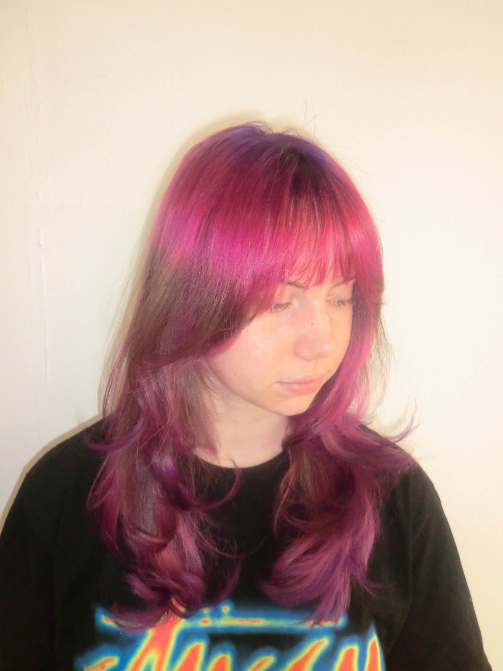 A front profile portrait of the same woman looking to the side, revealing a complex multi-tonal color transition. It features subtle "ghost roots" at the crown that blend from deep purple to crimson red and then into vivid pink. A long section of her natural dark blonde hair follows, before concluding with a vibrant pink dip-dye on the ends that gradients smoothly back into deep purple.