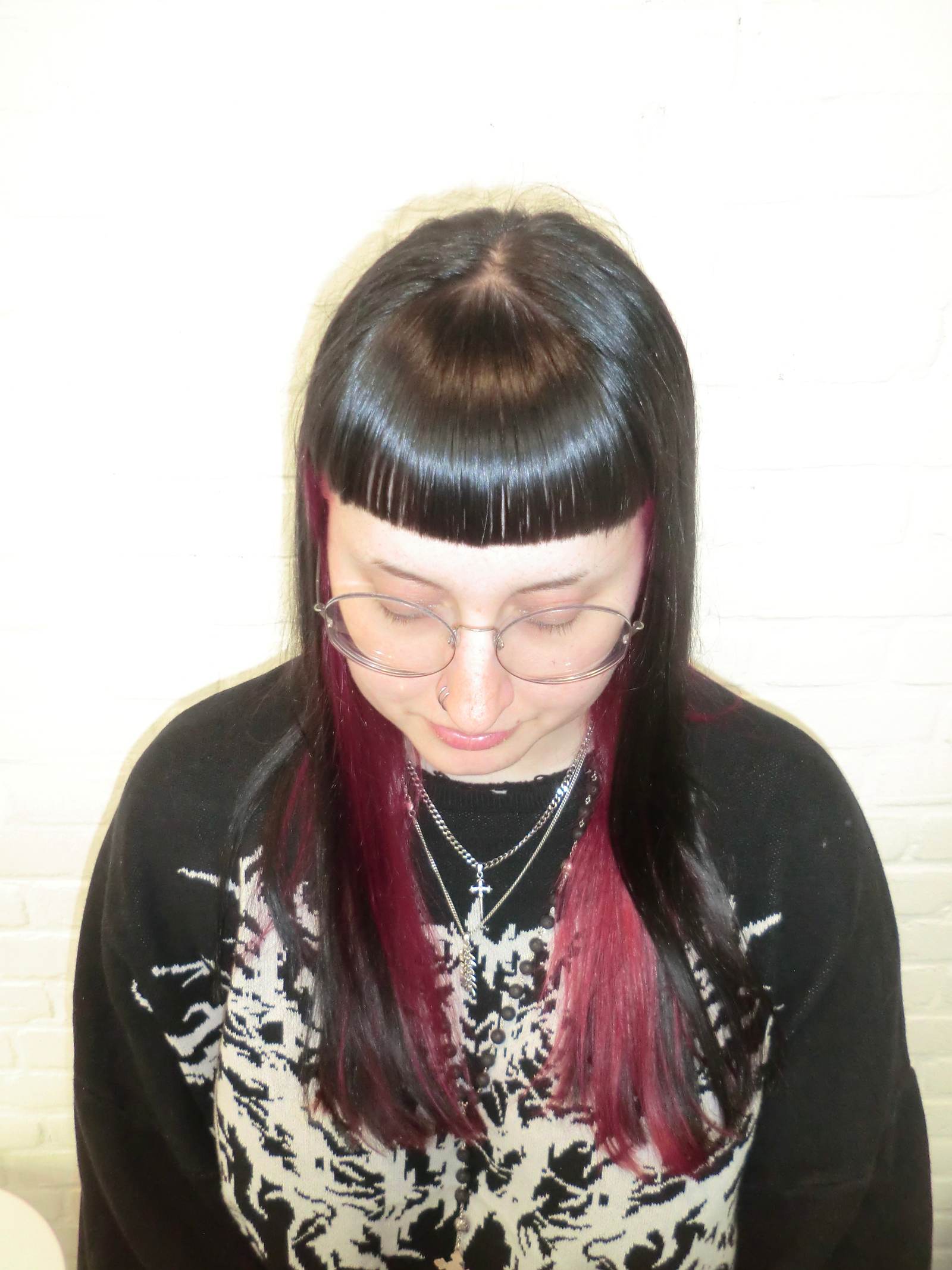A front profile taken from an upper angle, looking down at a high-contrast peekaboo hair transformation. The top layer of the hair is a deep jet-black, featuring full, straight-across bangs. From underneath, a vibrant, saturated bright red color is visible, creating a bold two-tone effect that peeks through the black layers and frames the face.