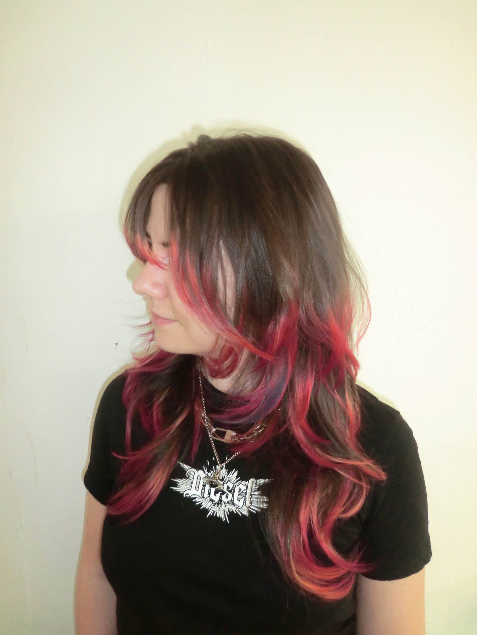 A side profile showcasing a vibrant "flame effect" dip-dye on a fresh layered cut. The natural brown hair transitions dramatically near the mid-lengths into a vivid, multi-tonal gradient of deep purple, fiery red, and bright neon orange at the tips. The texturized, face-framing layers emphasize the movement of the colors, making the ends look like flickering flames.