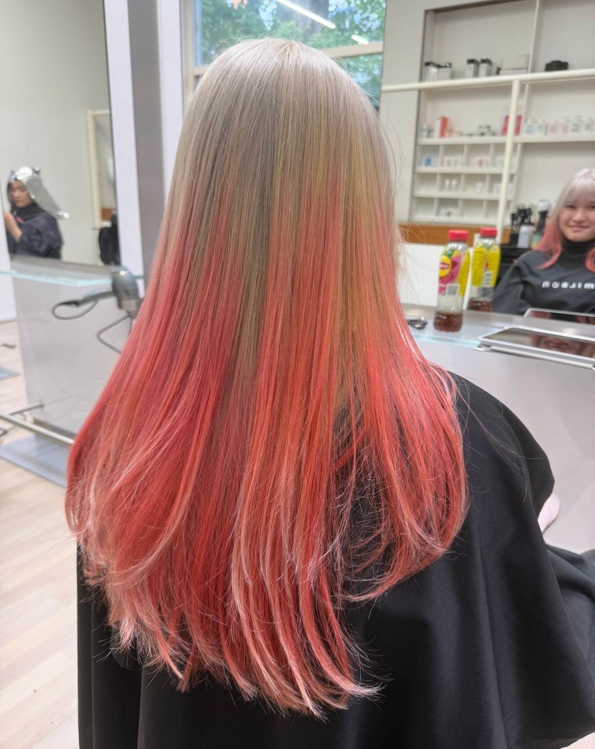 A back view of long, hair featuring a seamless color melt. The roots and mid-lengths are a cool, silverish blonde that transitions into a vibrant pink coral at the ends. The hair is styled that blend the icy metallic tones at the top with the warm, peachy-pink hues at the bottom.