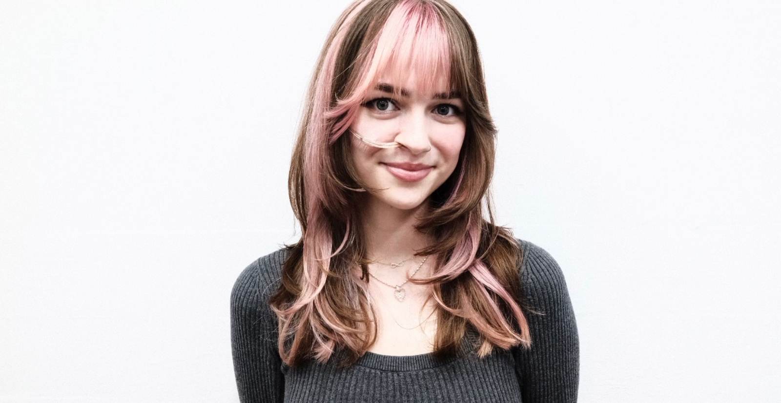 A front profile showing a bold hair transformation. The hair has been cut to chest-length and styled straight. It features large, chunky pink highlights that contrast sharply against a dark brunette base. The pink sections are particularly prominent around the face-framing layers and crown, creating a vibrant, color-blocked aesthetic.