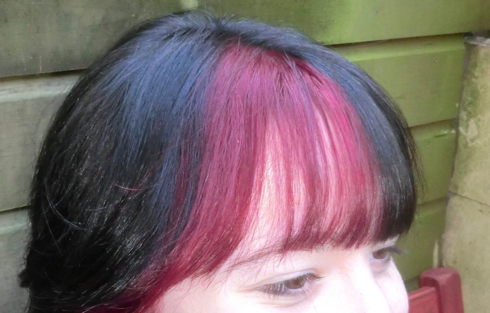 A close-up, front-view portrait focusing on a bold, high-contrast fringe. The hair is dyed a deep jet-black with thick, vibrant red streaks woven into the right side of the heavy, straight-across bangs. The red color is saturated and bright, creating a sharp graphic effect against the dark base. The bangs are cut thick to just above the eyebrows, framing the face and highlighting the vivid "peek-a-boo" red layer underneath.