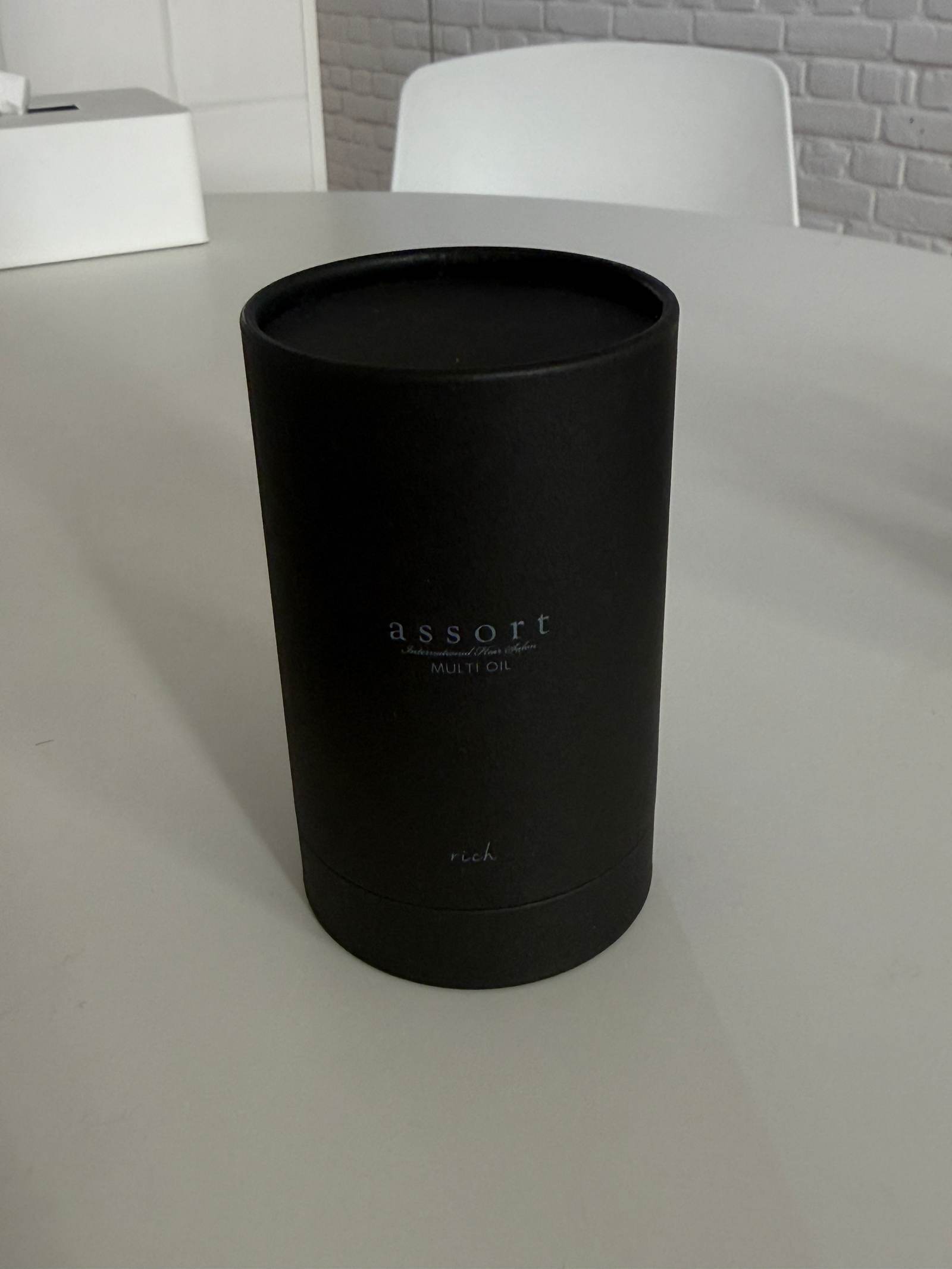 A professional shot of the Assort Multi Oil Rich in a tall, slender, black matte cylinder bottle. The bottle features a dropper-style cap and a minimalist white label. Its height is noticeably taller than the balm, conveying a premium, professional hair treatment aesthetic.