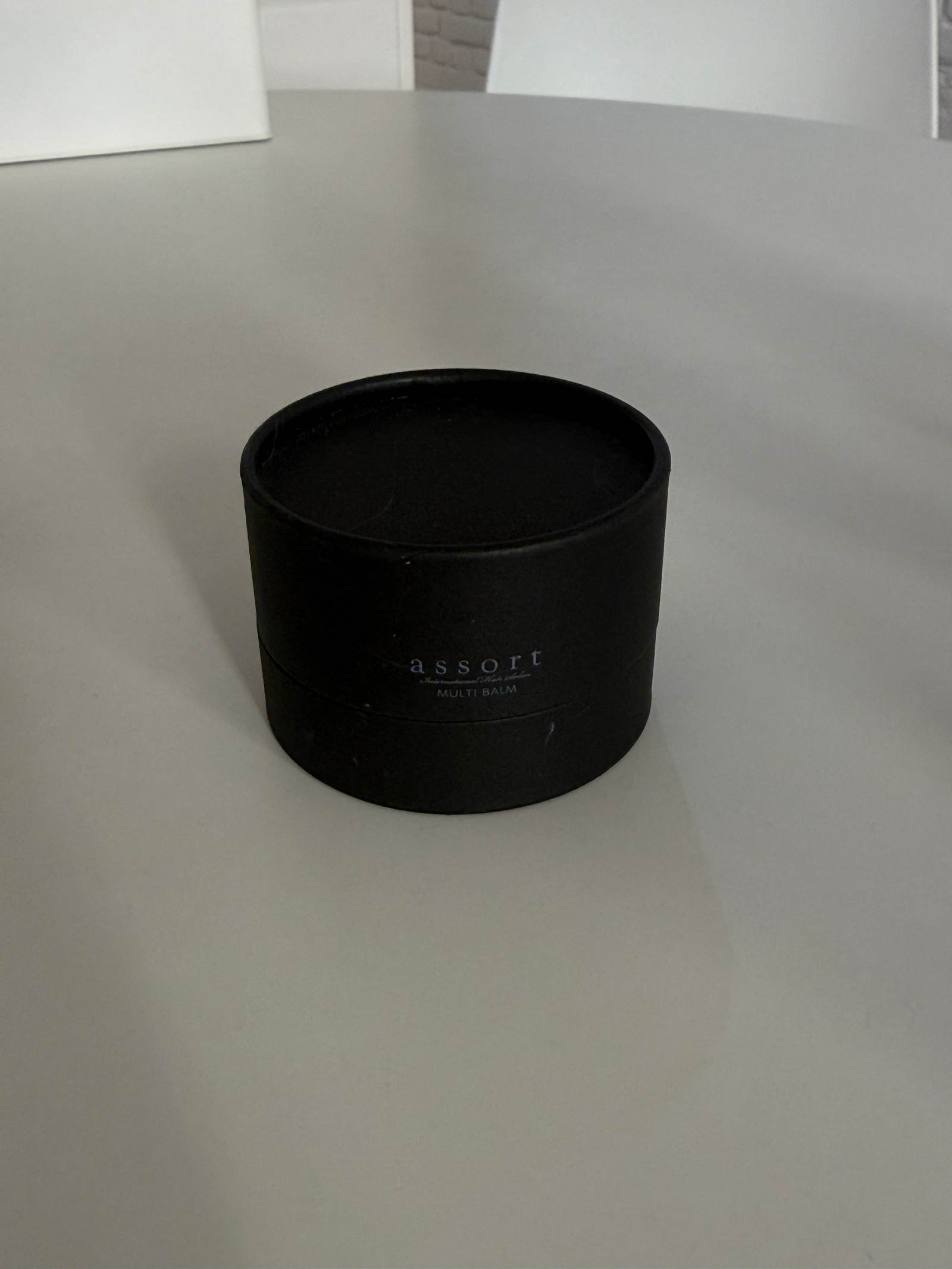 A close-up shot of the Assort Multi Balm in its signature packaging. The product is contained in a short, wide, black matte cylinder jar with a clean white label featuring minimalist black text. Its low-profile shape distinguishes it from the taller oil bottles in the collection.