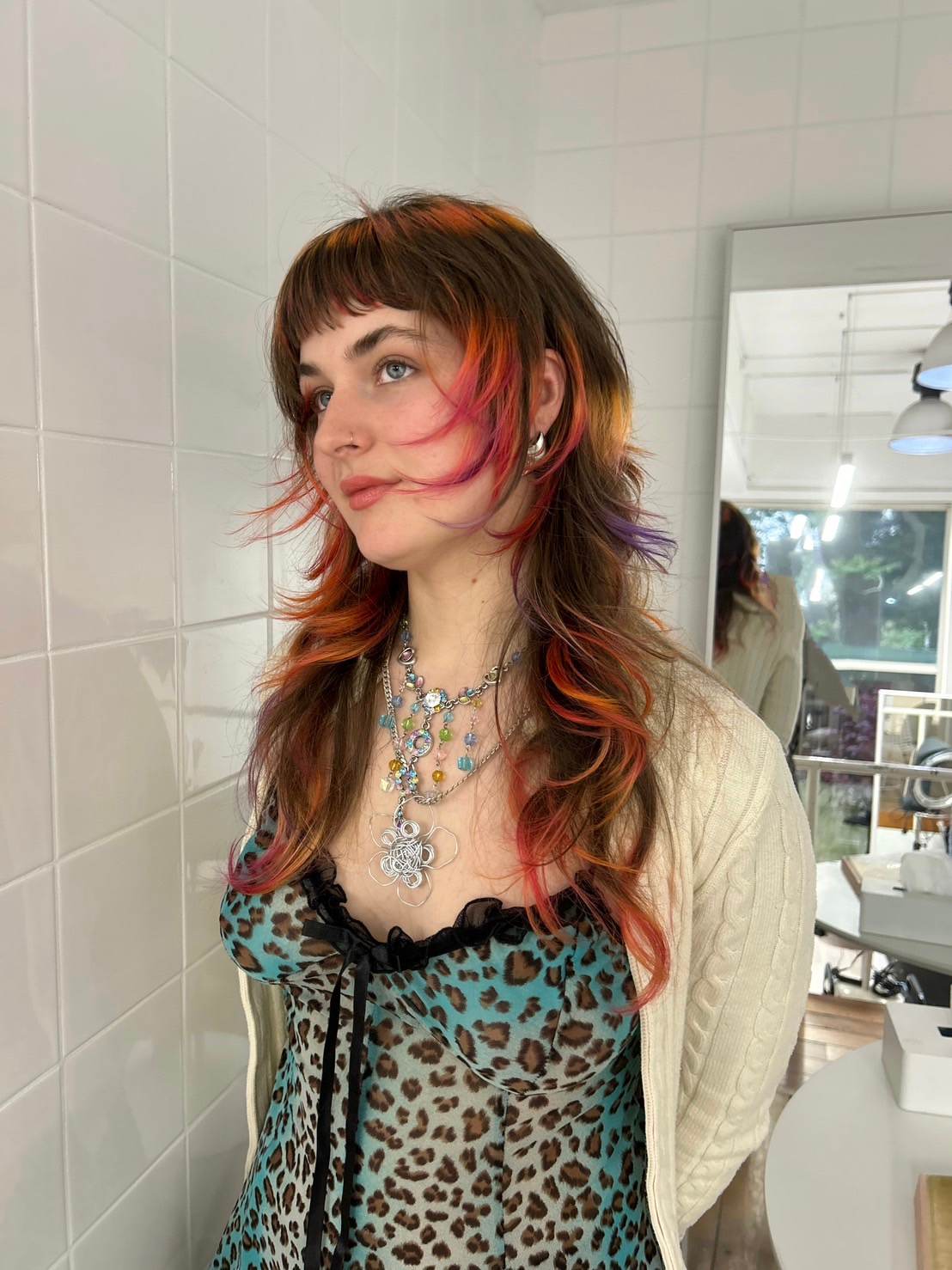 A front profile of a vibrant "sunset" hair transformation. The long, wavy brown-blonde hair features a "halo" of color around the face and crown, blending shades of soft purple, vivid pink, and warm orange into the blonde. The same sunset gradient is repeated at the ends in a dip-dye style, creating a colorful frame of pink and orange against the natural lengths.