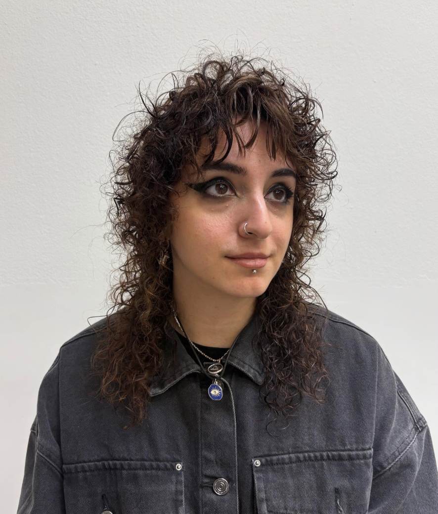 A front-facing portrait of a person with a shaggy, chest-length haircut and a fresh spiral perm. The hair is full of tight, springy ringlets that create significant volume and texture around the face. The shaggy layers and curly fringe frame the eyes and cheekbones, giving the style a modern, effortless "wolf cut" feel.