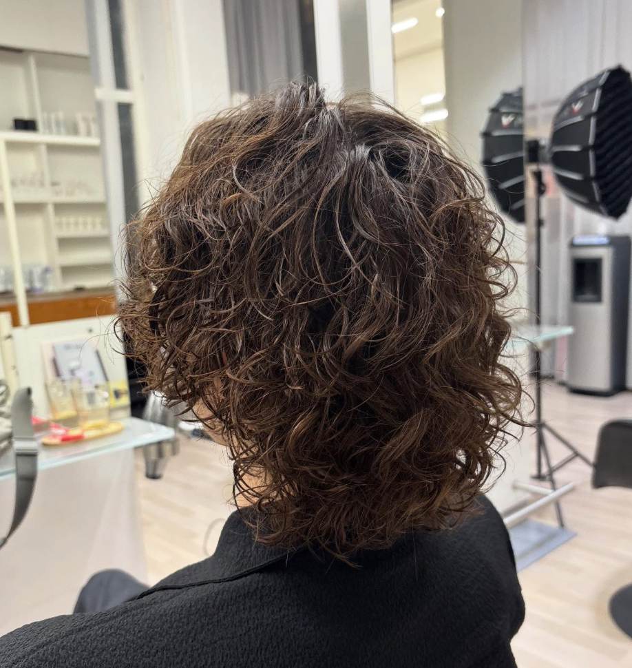 A back view of a textured haircut featuring a cold wave perm. The hair is styled into dense, well-defined curls that provide uniform volume and structure across the back of the head. The curls create a soft, rounded silhouette with significant lift, showcasing the consistent, springy texture characteristic of a cold wave treatment.