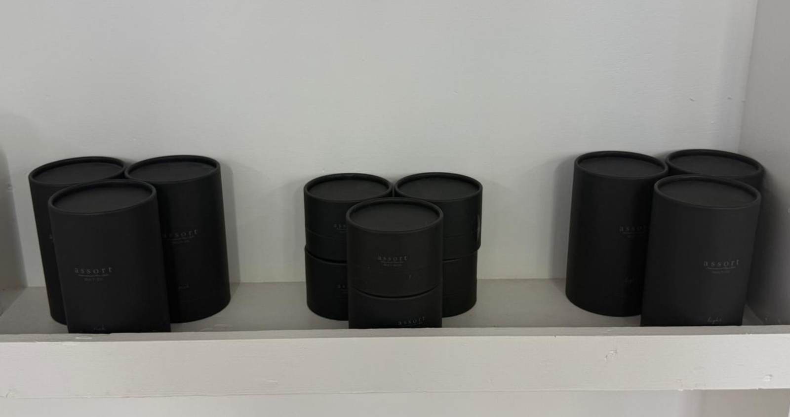 A lifestyle shot of the Assort product line arranged on a sleek salon shelf. The Assort Multi Balm, Multi Oil Rich, and Multi Oil Light are displayed together, showcasing their cohesive black matte cylinder packaging and clean white labels. The two oil bottles stand taller than the balm jar, creating a uniform and premium professional aesthetic.