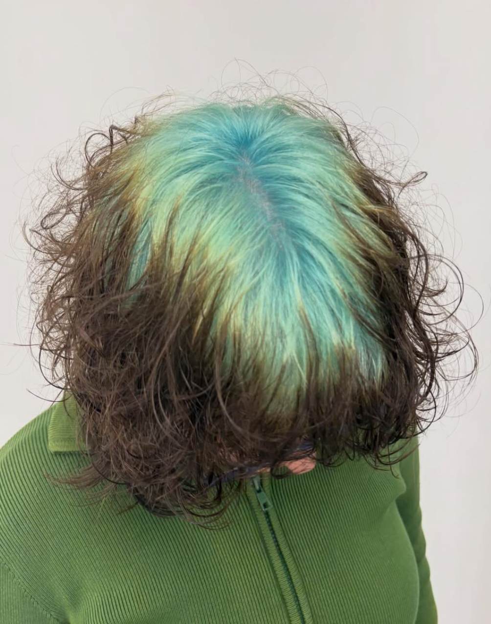 A front view of a short bixie haircut where the client is looking down, causing the top layer of natural brown hair to cascade slightly forward. This position reveals a prominent, contrasting band of vibrant teal-mint green color applied directly at the roots across the crown and parting line, characteristic of the "ghost root" technique.