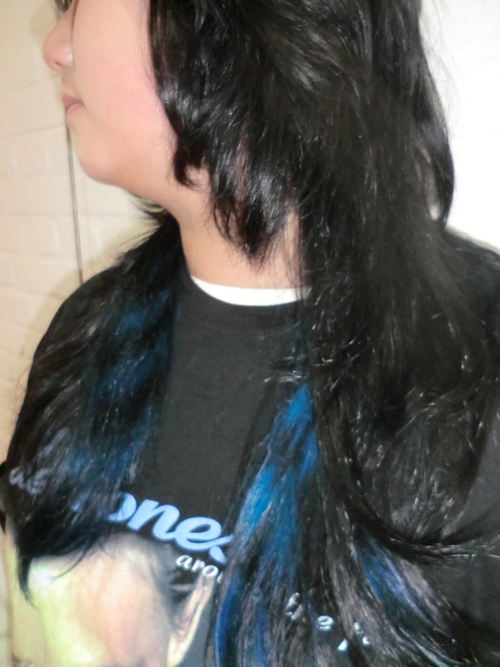 A side profile of a person with long, black hair. The top layer of hair is pulled back, revealing a vibrant, electric blue "underlight" color underneath. The contrasting blue strands flow alongside the natural dark hair, showcasing the hidden color technique.