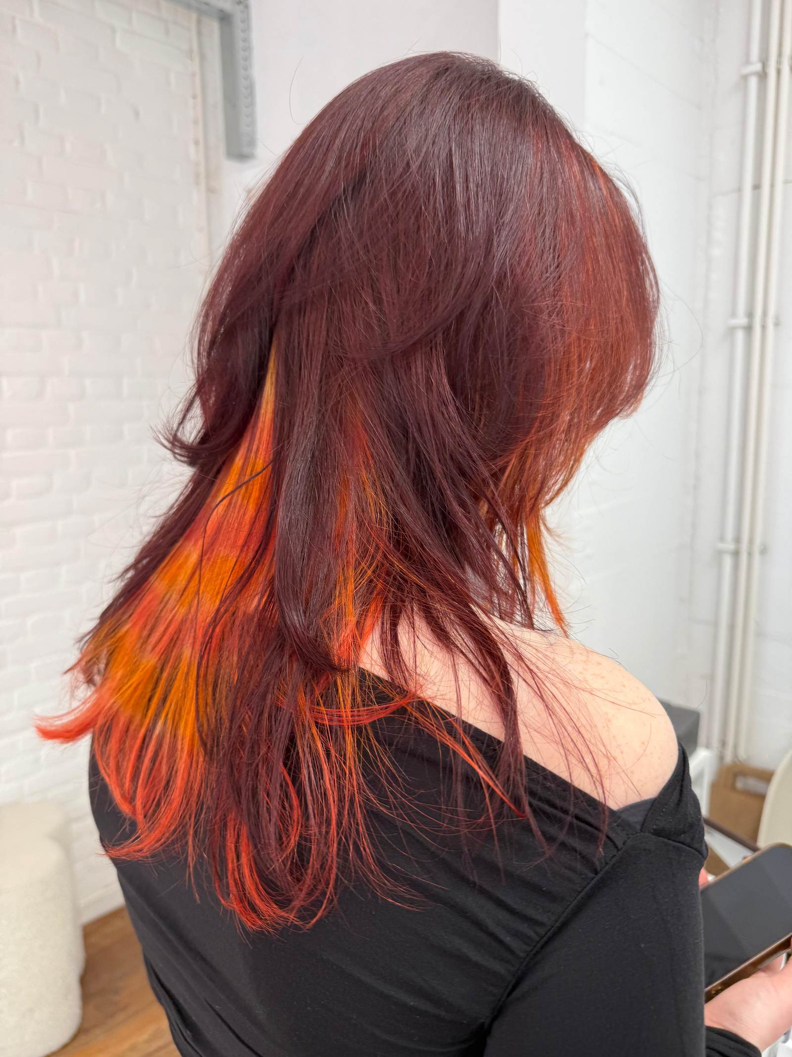 A back view of the vibrant red hairstyle. The top layer is a solid, intense red, which lifts to reveal a hidden "peekaboo" underlayer of bright neon orange. The sides of the hair show the continuation of the "raccoon tail" striped pattern, creating a high-contrast, multi-dimensional look that pops against the dark red surface.
