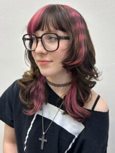 A portrait of a person with a layered, shoulder-length brown "wolf cut" featuring vibrant pink accents. The hair has a bright pink "money piece" fringe and a unique checkered or "pixelated" pink pattern dyed into the side layers.