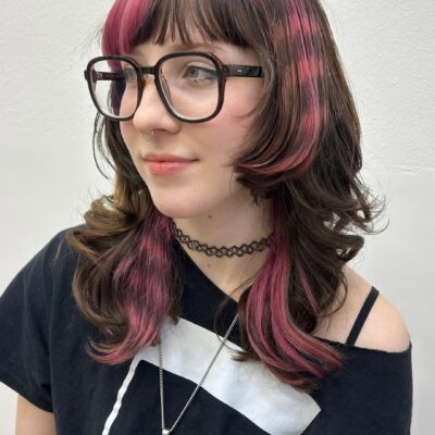 A portrait of a person with a layered, shoulder-length brown "wolf cut" featuring vibrant pink accents. The hair has a bright pink "money piece" fringe and a unique checkered or "pixelated" pink pattern dyed into the side layers.