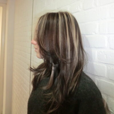 A profile view of a person with long, dark brown hair featuring bold, chunky blonde highlights. The hair is styled in loose waves and layered around the face. They are wearing a dark charcoal sweater and standing against a white brick wall.