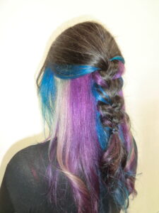 A back view of long, straight hair featuring a vibrant "peek-a-boo" color style with layers of electric blue and bright purple underneath a dark brunette top layer. The hair is partially styled in a loose French braid down the center, which pulls the blue and purple tones through to the surface, creating a woven, multi-colored effect against the dark hair.