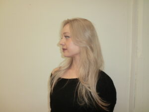 A side profile showing the layered cut of the platinum blonde hair. The long layers curve gently around the face and shoulders, highlighting the uniform, cool-toned color.