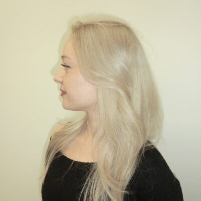 A side profile showing the layered cut of the platinum blonde hair. The long layers curve gently around the face and shoulders, highlighting the uniform, cool-toned color.