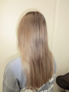 A back view of long, even-toned hair in a warm "dirty blonde" shade. The color is a consistent blend of honey and light brown tones with no visible regrowth. The hair has a smooth, straight texture and a clean, blunt-cut hemline that falls to the mid-back, serving as the base for a future color or texture transformation.