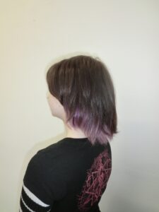 A portrait of a person with shoulder-length brown hair and straight-across bangs. The style features a "peek-a-boo" effect with a hidden layer of vibrant purple hair underneath the natural brown top section.