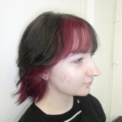 A portrait of a person showcasing a bold color transformation to jet-black hair. The look includes thicker, full bangs with bright red streaks concentrated on the right side, along with a hidden layer of vivid red hair throughout the underneath sections.