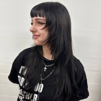 A portrait of a person with long black hair that has been cut into textured layers and styled with voluminous waves. The look features soft, face-framing "curtain" bangs and shorter layers throughout the lengths to add movement and body to the style.