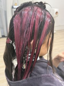 A close-up, back-view shot of a hair coloring process in a salon. Vibrant magenta-pink hair dye is being applied in thick vertical sections to dark brown hair. Some lower sections are wrapped in silver foil to keep them separate, while pink-saturated strands are visible against the dark uncolored hair.