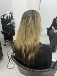 A back view of long hair showing significant root regrowth and a faded color treatment. The top section features several inches of natural dark espresso roots transitioning into a muted, warm-toned blonde. The bottom ends show the remains of a black dip dye that has faded into a soft, smoky charcoal grey, with the hair tapering into natural, unstyled waves.