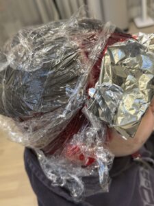 A close-up, back-view shot of a professional hair coloring session in progress. Vibrant red hair dye is being applied in thick sections to dark hair, while other sections are separated and held in place by silver foils. The foils are concentrated around the nape and the right side of the head, indicating where the hidden layers and specific streaks are being processed. The person is wearing a black salon cape to protect their clothing during the application.