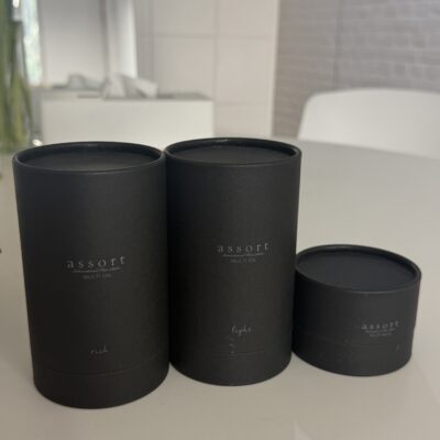 A professional product shot of three Assort hair care items standing side-by-side against a clean background. From left to right: the Assort Multi Balm in a short, wide black matte cylinder jar with a minimalist white label, the Assort Multi Oil Rich in a taller black matte cylinder bottle with a dropper top, and the Assort Multi Oil Light in an identical tall black matte bottle. The heights create a stepped look, with the oils standing taller than the balm.