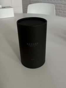 A professional shot of the Assort Multi Oil Rich in a tall, slender, black matte cylinder bottle. The bottle features a dropper-style cap and a minimalist white label. Its height is noticeably taller than the balm, conveying a premium, professional hair treatment aesthetic.