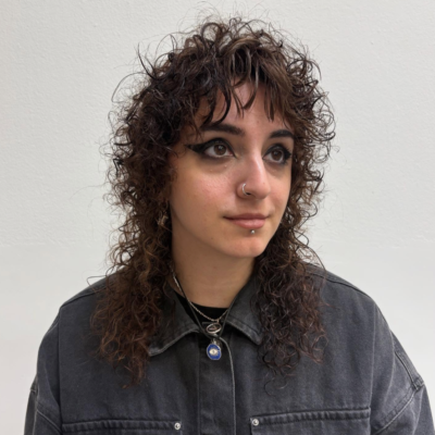 A front-facing portrait of a person with a shaggy, chest-length haircut and a fresh spiral perm. The hair is full of tight, springy ringlets that create significant volume and texture around the face. The shaggy layers and curly fringe frame the eyes and cheekbones, giving the style a modern, effortless "wolf cut" feel.