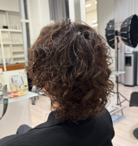 A back view of a textured haircut featuring a cold wave perm. The hair is styled into dense, well-defined curls that provide uniform volume and structure across the back of the head. The curls create a soft, rounded silhouette with significant lift, showcasing the consistent, springy texture characteristic of a cold wave treatment.