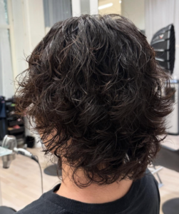 A back view of a textured haircut featuring a cold wave perm. The hair is styled into dense, well-defined curls that provide uniform volume and structure across the back of the head. The curls create a soft, rounded silhouette with significant lift, showcasing the consistent, springy texture characteristic of a cold wave treatment.