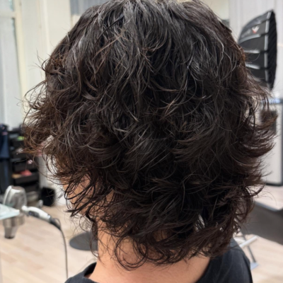 A back view of a textured haircut featuring a cold wave perm. The hair is styled into dense, well-defined curls that provide uniform volume and structure across the back of the head. The curls create a soft, rounded silhouette with significant lift, showcasing the consistent, springy texture characteristic of a cold wave treatment.
