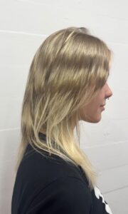 A side profile showing the completed hairstyle from the previous process photo. The hair is a bright, creamy blonde with subtle darker lowlights for dimension. It is styled with a soft, layered cut and face-framing fringe that creates a modern, effortless look against a white wall.