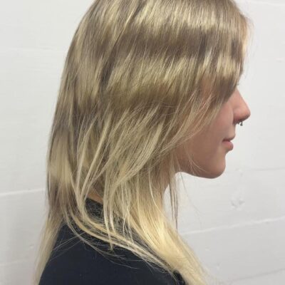 A side profile showing the completed hairstyle from the previous process photo. The hair is a bright, creamy blonde with subtle darker lowlights for dimension. It is styled with a soft, layered cut and face-framing fringe that creates a modern, effortless look against a white wall.