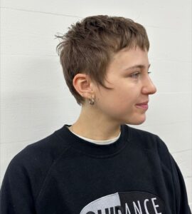 A side profile of a classic pixie cut. The hair is cropped short around the sides and back, with slightly more length and texture on top to create a clean, tapered silhouette.