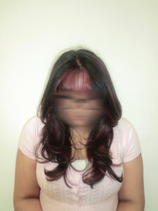 A front profile of a person with long, wavy black hair featuring a high-contrast pink transformation. The full, straight-across bangs are dyed a vibrant magenta pink, which continues into "peekaboo" sections underneath the black top layers. The pink strands frame the face and peek through the dark curls, creating a bold and edgy two-tone look.