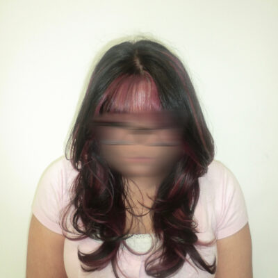 A front profile of a person with long, wavy black hair featuring a high-contrast pink transformation. The full, straight-across bangs are dyed a vibrant magenta pink, which continues into "peekaboo" sections underneath the black top layers. The pink strands frame the face and peek through the dark curls, creating a bold and edgy two-tone look.