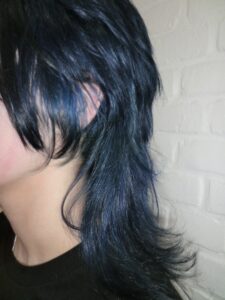 A side profile view of a girl with a shoulder-length layered mullet. The hair is a vibrant electric blue from the roots through the mid-lengths, ending in a sharp black dip dye at the tips. The cut features choppy, textured layers with a shorter top and longer back, showcasing a modern, edgy silhouette.