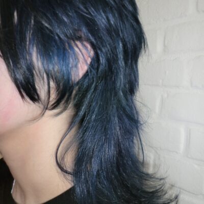 A side profile view of a girl with a shoulder-length layered mullet. The hair is a vibrant electric blue from the roots through the mid-lengths, ending in a sharp black dip dye at the tips. The cut features choppy, textured layers with a shorter top and longer back, showcasing a modern, edgy silhouette.