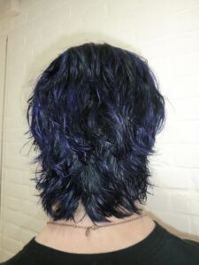 A back view of a male with a short, textured haircut featuring "ghost roots." The hair is dyed a deep natural black at the roots and crown, transitioning sharply into a vibrant electric blue at the ends. The cut is short around the sides and nape, with slightly more length on top to show the contrast between the dark roots and the bright blue tips.