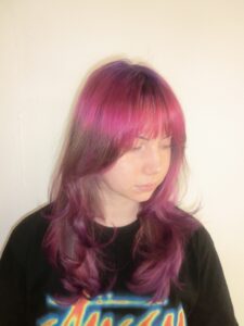 A front profile portrait of the same woman looking to the side, revealing a complex multi-tonal color transition. It features subtle "ghost roots" at the crown that blend from deep purple to crimson red and then into vivid pink. A long section of her natural dark blonde hair follows, before concluding with a vibrant pink dip-dye on the ends that gradients smoothly back into deep purple.