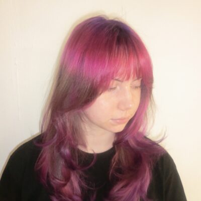A front profile portrait of the same woman looking to the side, revealing a complex multi-tonal color transition. It features subtle "ghost roots" at the crown that blend from deep purple to crimson red and then into vivid pink. A long section of her natural dark blonde hair follows, before concluding with a vibrant pink dip-dye on the ends that gradients smoothly back into deep purple.