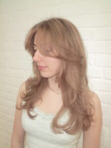 A side profile view of a medium-length hairstyle featuring a soft "beige-ginger" color. The look blends warm copper tones with muted, sandy-beige highlights to create a multi-dimensional, natural-looking finish. The hair is styled in soft waves, showcasing how the lighter beige pieces catch the light against the warm ginger base.