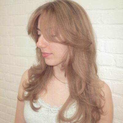 A side profile view of a medium-length hairstyle featuring a soft "beige-ginger" color. The look blends warm copper tones with muted, sandy-beige highlights to create a multi-dimensional, natural-looking finish. The hair is styled in soft waves, showcasing how the lighter beige pieces catch the light against the warm ginger base.