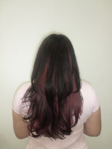 Alt Text: A front profile of a person with long, wavy black hair featuring a high-contrast pink transformation. The full, straight-across bangs are dyed a vibrant magenta pink, which continues into "peekaboo" sections underneath the black top layers. The pink strands frame the face and peek through the dark curls, creating a bold and edgy two-tone look.