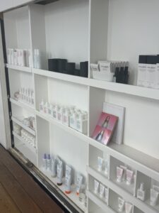 A wide-angle shot of a minimalist wooden salon shelf displaying an organized variety of professional hair care products. The shelf includes various bottles of shampoo, conditioner, styling oils, and tubs of hair balm in neutral-toned packaging. The products are arranged neatly, creating a clean and professional retail display.